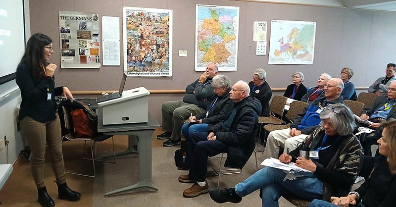 Instructor Pamela Ahern presents to a seated group of adult learners at the Osher Lifelong Learning Institute. Participants are gathered in a classroom, listening to the presentation, with maps and posters displayed on the wall behind the instructor.