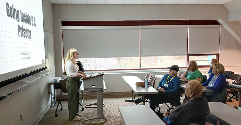 MacKenzie Niness teaches a class at the Osher Lifelong Learning Institute, standing beside a presentation screen while adult learners seated at tables follow along in a classroom setting.